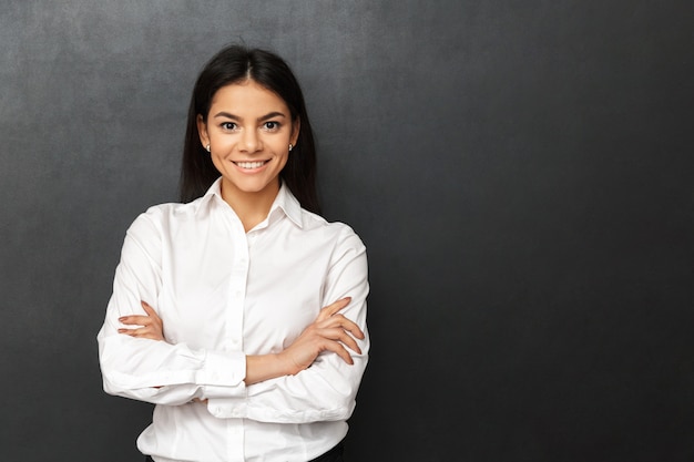 Portrait de femme de race blanche aux longs cheveux bruns portant des vêtements de bureau souriant tout en posant les bras croisés, isolé sur mur gris foncé