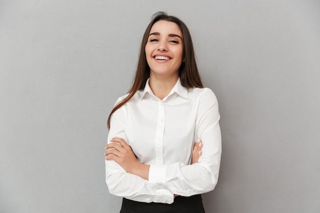 Portrait de femme de race blanche avec de beaux longs cheveux bruns en affaires porter souriant et gardant les bras croisés, isolé sur mur gris