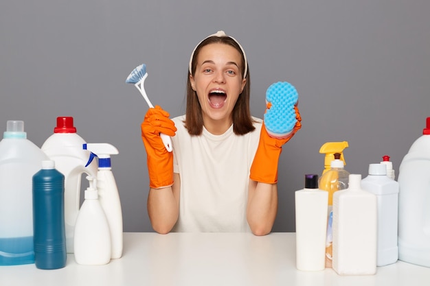 Portrait d'une femme ravie excitée porte des gants orange bandeau et un t-shirt décontracté assis sur le lieu de travail utilise différents détergents tenant une brosse et un plumeau pp dans les mains isolés sur fond gris