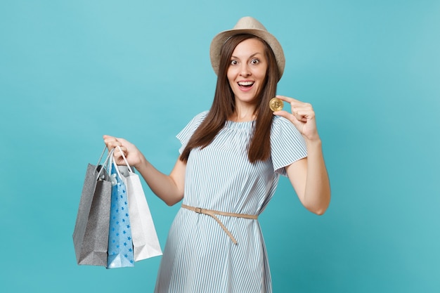 Portrait de femme en robe d'été, chapeau de paille tenant des paquets de sacs avec achats après le shopping, bitcoin, pièce de métal de couleur dorée isolée sur fond bleu pastel. Copiez l'espace pour la publicité.