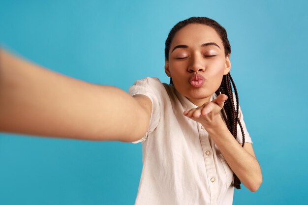 Portrait d'une femme romantique avec des dreadlocks faisant selfie en envoyant des baisers aériens avec les yeux fermés point de vue photo portant une chemise blanche Prise de vue en studio intérieure isolée sur fond bleu