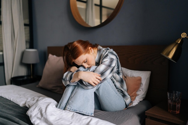 Portrait d'une femme rousse millénaire triste bouleversée assise sur son lit embrasser les genoux perdus dans de mauvaises pensées pessimistes souffrir seule Une jeune femme stressée se sent seule