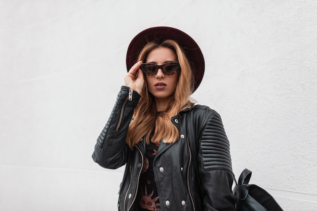 Portrait d'une femme rousse sexy jeune hipster dans un élégant chapeau à lunettes de soleil sombres dans une veste en cuir à la mode avec un sac à dos