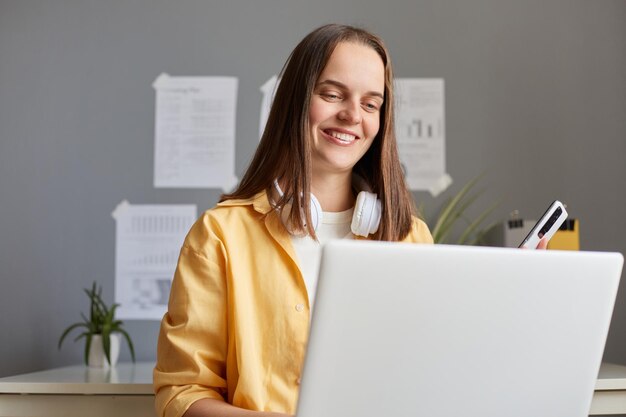 Portrait d'une femme séduisante aux cheveux bruns travaillant sur un ordinateur portable au bureau et tenant un téléphone intelligent dans les mains regardant le moniteur avec le sourire étant heureuse de son travail