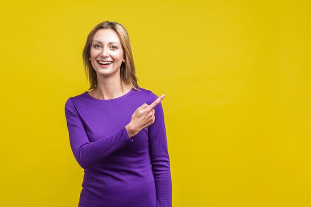 Portrait d'une femme séduisante et joyeuse dans une élégante robe violette serrée, debout, pointant du doigt l'espace de copie vide, place pour la publicité. studio d'intérieur tourné isolé sur fond jaune