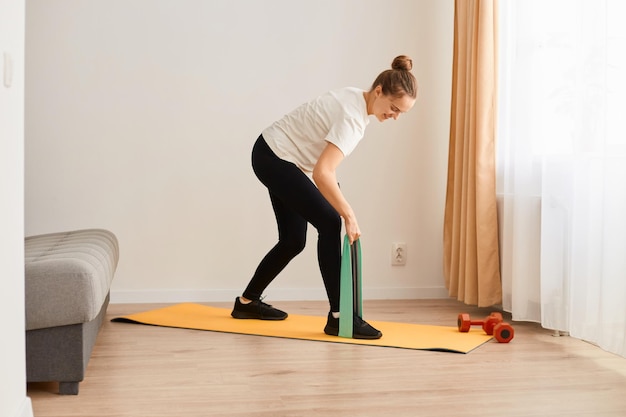 Portrait d'une femme séduisante mince portant un t-shirt blanc et des leggins noirs faisant des exercices de sport à la maison debout avec le haut du corps plié et tenant une bande de résistance pour l'entraînement