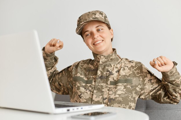 Photo portrait d'une femme soldat souriante et satisfaite portant un uniforme de camouflage et une casquette terminant son travail en ligne exprimant le bonheur assis et étirant les bras