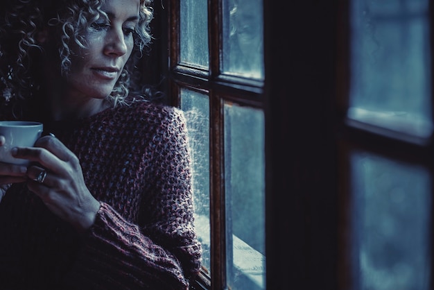 Portrait de femme solitaire aux tons bleus regardant par les fenêtres à la maison en buvant du thé la nuit. Concept de la tristesse et de la maladie de l'insomnie. Concept unique de personnes de sexe féminin adultes attrayantes