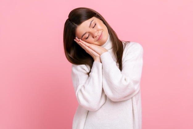 Portrait d'une femme souriante à l'air agréable, tenant la tête sur la main, gardant les yeux fermés, paresse, portant un pull blanc de style décontracté. Tourné en studio intérieur isolé sur fond rose.