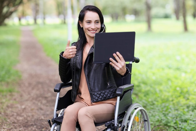 Portrait de femme souriante assise en fauteuil roulant dans le parc et montrant le pouce vers le haut sur la tablette de