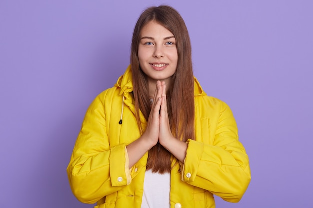 Portrait de femme souriante aux cheveux bruns habille la veste jaune, gardant les paumes ensemble et priant, regardant la caméra avec un sourire agréable, isolé sur fond lilas.