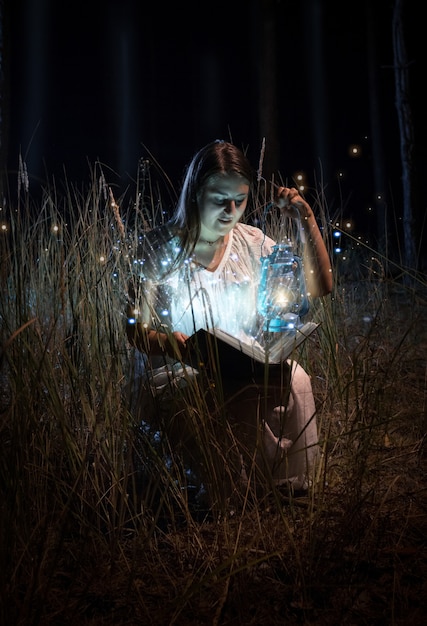 Portrait de femme souriante en chemise de nuit assise sur le terrain la nuit et livre de lecture