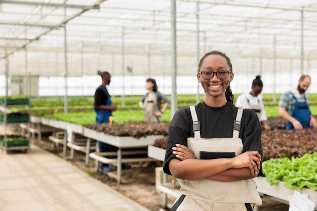 Portrait d'une femme souriante dans une serre moderne avec des ouvriers prenant soin des semis de salades et de plantes vertes. Ouvrier agricole afro-américain posant heureux dans une ferme de légumes biologiques.