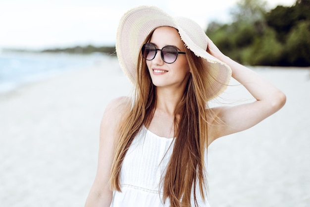 Portrait d'une femme souriante heureuse dans le bonheur gratuit sur la plage de l'océan debout avec un chapeau et des lunettes de soleil. Un modèle féminin dans une robe d'été blanche profitant de la nature pendant les vacances de voyage vacances outd