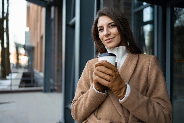 Portrait d'une femme souriante en manteau avec une tasse de café près de l'immeuble de bureaux