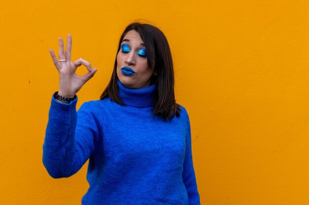 portrait d'une femme souriante avec maquillage et robe bleue faisant le symbole OK avec la main sur fond jaune. concept de féminisme, journée de la femme, 8 mars.