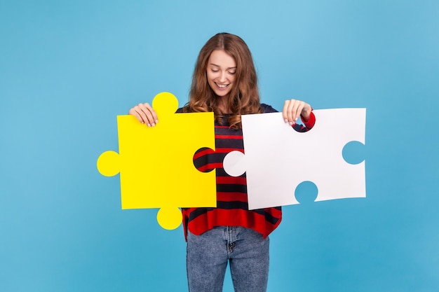 Portrait d'une femme souriante portant un pull rayé de style décontracté, rejoignant deux pièces de puzzle, métaphore de la connexion d'unité, solution complète. Studio intérieur tourné isolé sur fond bleu.