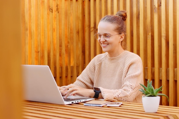 Portrait d'une femme souriante ravie et ravie portant un pull beige travaillant sur un ordinateur portable au bureau contre un mur en bois tapant sur un ordinateur portable avec une expression faciale heureuse