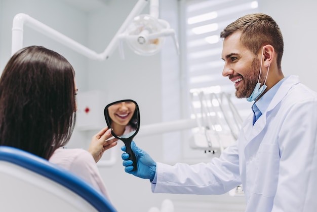 Portrait d'une femme souriante regardant dans le miroir en train de vérifier les dents après une intervention dentaire en clinique