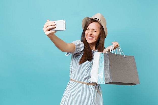 Portrait femme souriante en robe d'été, chapeau de paille tenant des paquets de sacs avec des achats après le shopping en faisant un selfie photo sur téléphone portable isolé sur fond bleu pastel. Espace de copie pour la publicité