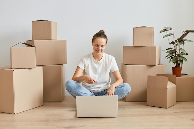 Portrait d'une femme souriante et satisfaite portant un t-shirt blanc et un jean assis sur le sol entouré de boîtes en carton et travaillant sur un ordinateur portable pointant vers l'écran de l'ordinateur portable avec le doigt