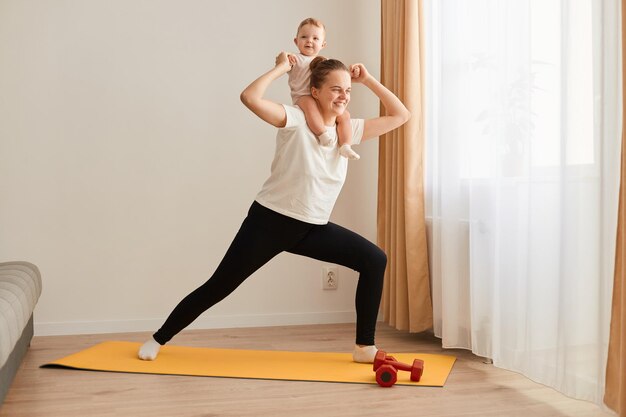 Portrait d'une femme sportive faisant du yoga sur un tapis de yoga à la maison avec sa petite fille sur son épaule mode de vie sain exercices de sport de remise en forme près de la fenêtre i salon