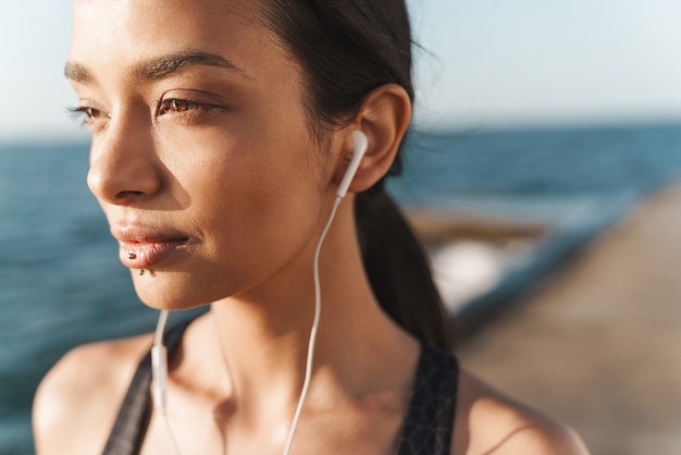 Portrait d'une femme sportive forte et heureuse à l'extérieur sur la plage le matin posant en écoutant de la musique avec des écouteurs.