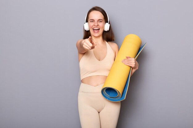 Portrait d'une femme sportive en tenue de sport beige porte des poses de karemat roulées isolées sur fond gris pointant vers la caméra écoutant de la musique