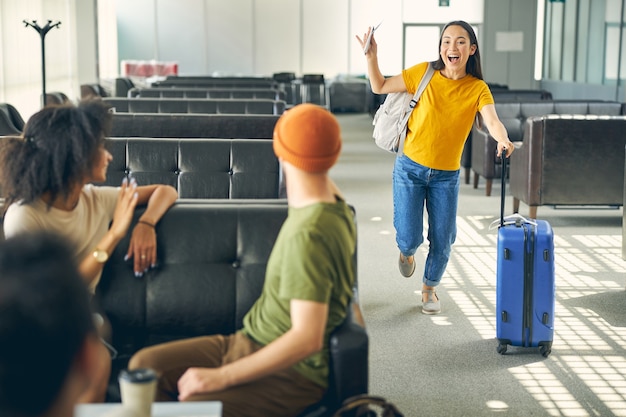 Portrait d'une femme sympathique avec une valise bleue et un sac à dos gris courant vers le meilleur groupe de touristes à l'aéroport