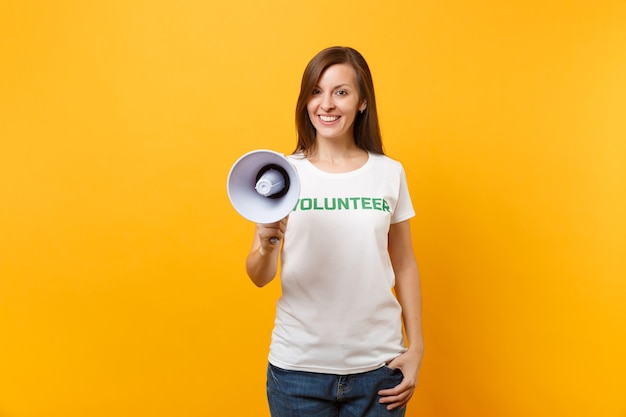 Portrait de femme en t-shirt blanc écrit inscription verte titre volontaire crier dans un mégaphone de sonorisation isolé sur fond jaune. Aide d'assistance gratuite volontaire, concept de travail de grâce de charité.