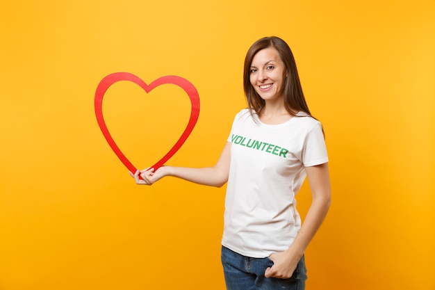 Portrait de femme en t-shirt blanc avec inscription écrite titre vert bénévole tenir grand coeur en bois rouge isolé sur fond jaune. Aide d'assistance gratuite volontaire, concept de travail de grâce de charité.