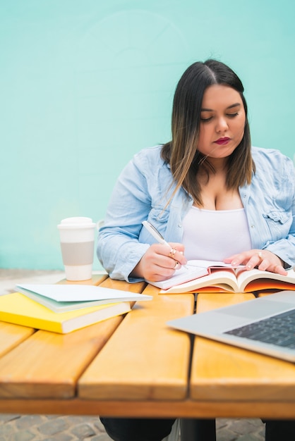 Portrait de femme de taille plus jeune étudiant avec ordinateur portable et livres alors qu'il était assis à l'extérieur au café.