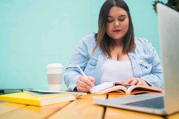 Portrait de femme de taille plus jeune étudiant avec ordinateur portable et livres alors qu'il était assis à l'extérieur au café.