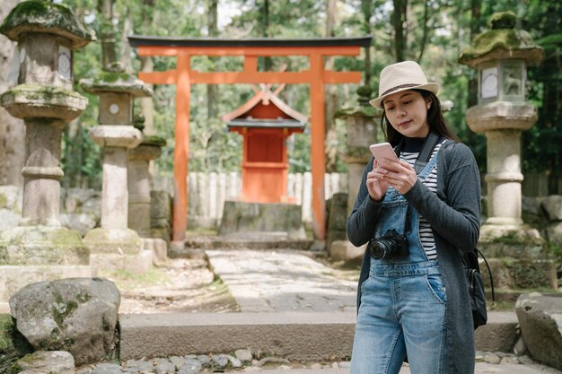 portrait femme taïwanaise debout devant le torii et le sanctuaire pour utiliser le téléphone. un joli routard asiatique portant une caméra recherche des informations sur un smarphone.