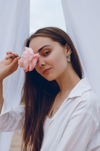 Portrait d'une femme tendre couvrant les yeux avec une fleur rose rose et regardant la caméra à l'extérieur