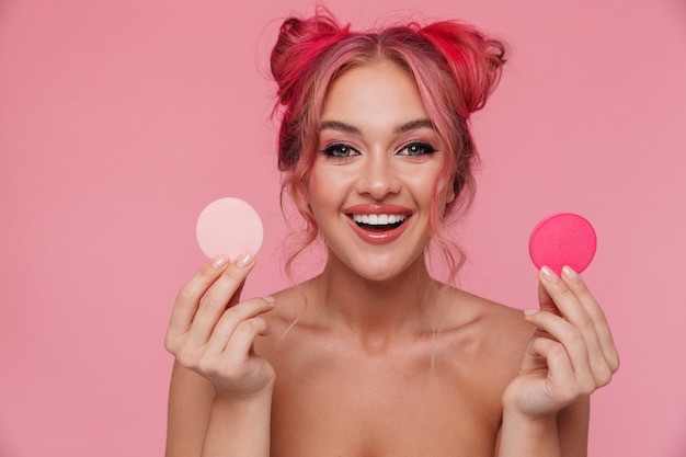 Portrait de femme torse nu joyeuse avec une coiffure colorée tenant des éponges de maquillage