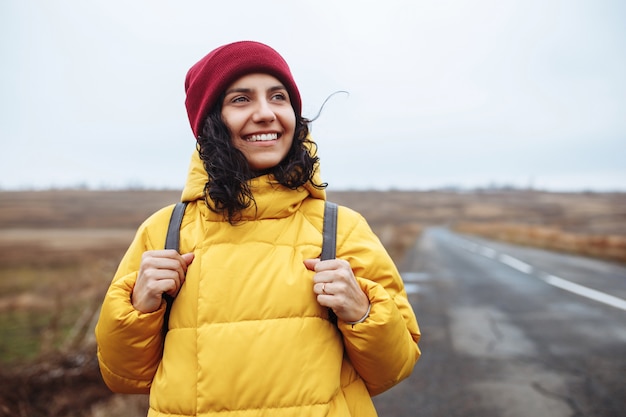 Portrait d'une femme touriste avec un sac à dos portant une veste jaune et un chapeau rouge se dresse sur la route.