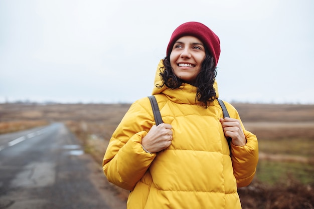 Portrait d'une femme touriste avec un sac à dos portant une veste jaune et un chapeau rouge se dresse sur la route.