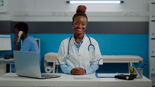 Portrait d'une femme travaillant comme médecin au bureau d'une clinique de soins de santé. Médecin portant une blouse blanche et un stéthoscope assis au bureau dans une armoire médicale souriant et regardant la caméra