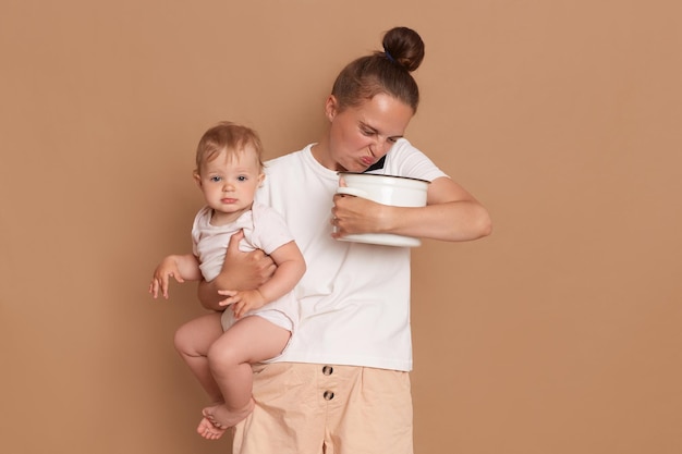 Portrait d'une femme triste portant une chemise blanche de style décontracté tenant dans les bras sa petite fille et sentant la nourriture gâtée dans un pot tout en parlant au téléphone portable isolé sur fond marron