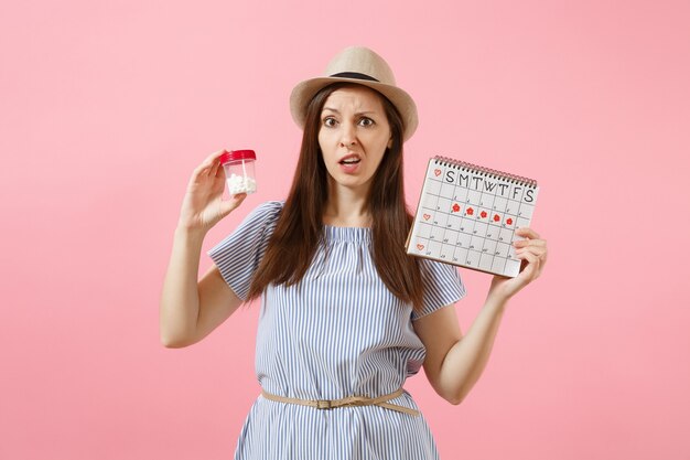 Portrait d'une femme triste en robe bleue tenant une bouteille blanche avec des pilules, calendrier des périodes féminines, vérifiant les jours de menstruation isolés sur fond. Soins médicaux, concept gynécologique. Espace de copie.