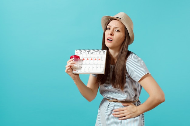 Portrait d'une femme triste en robe tenant une bouteille blanche avec des pilules, calendrier des périodes féminines, vérifiant les jours de menstruation isolés sur fond bleu. Soins médicaux, concept gynécologique. Espace de copie.