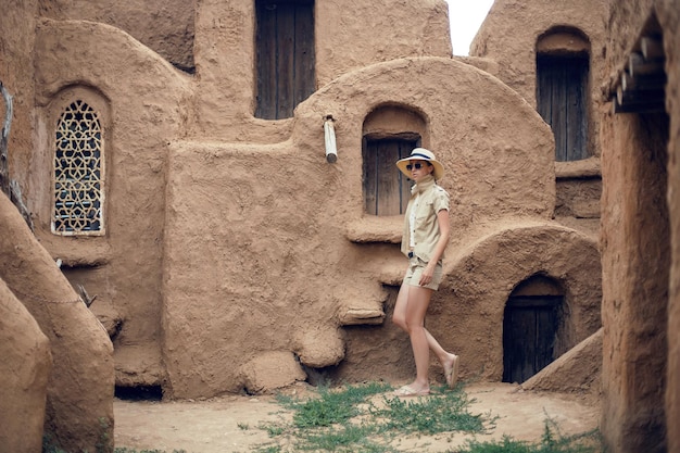 Portrait d'une femme voyageuse dans un chapeau et des lunettes de soleil se dresse dans une ville orientale abandonnée faite d'argile en short et veste de sable court d'été