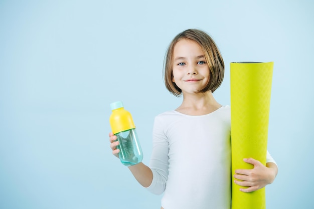 Portrait d'une fille de 8 ans dans un justaucorps blanc tenant une bouteille d'eau et un tapis de yoga