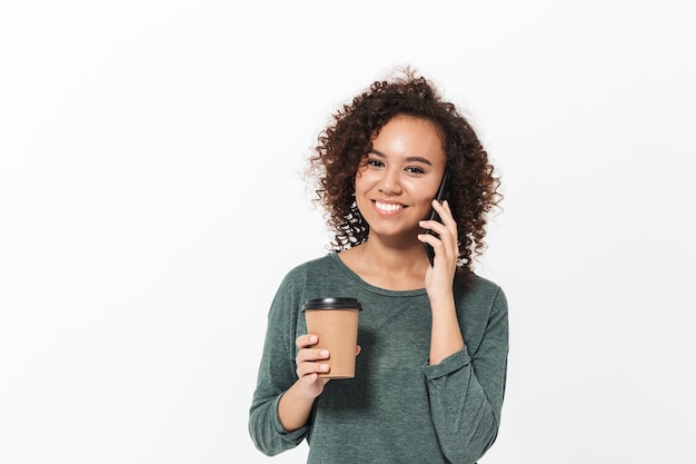 Portrait d'une fille africaine décontractée assez joyeuse debout isolée sur un mur blanc, parlant au téléphone portable, buvant du café à emporter