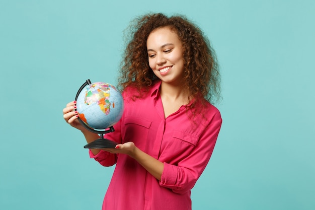 Portrait d'une fille africaine souriante dans des vêtements décontractés tenant dans les mains un globe terrestre isolé sur fond bleu turquoise en studio. Les gens émotions sincères, concept de style de vie. Maquette de l'espace de copie.