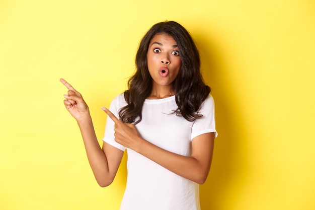Portrait d'une fille afro-américaine surprise en t-shirt blanc, l'air étonné et pointant du doigt dans le coin supérieur gauche, montrant votre logo ou votre bannière promotionnelle sur fond jaune.