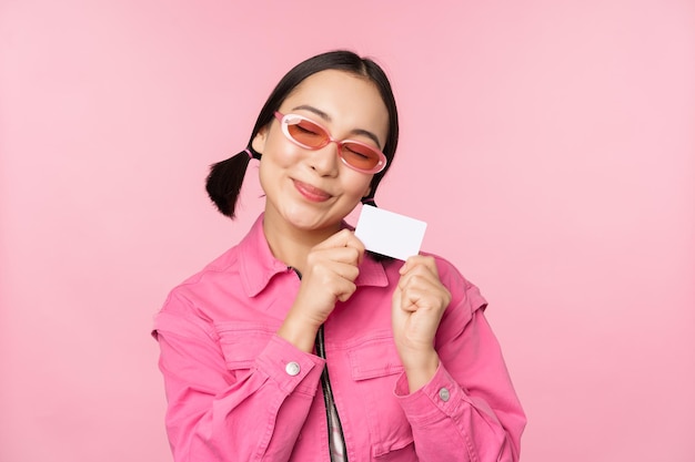 Portrait d'une fille asiatique moderne et élégante montre une carte de crédit à prix réduit et a l'air heureux de payer le concept sans contact de shopping debout sur l'espace de copie de fond rose