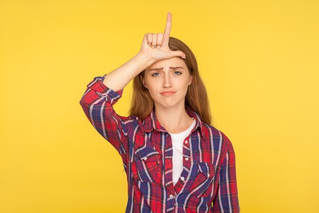 Portrait d'une fille au gingembre malchanceuse frustrée en chemise décontractée montrant le geste du perdant L signe du doigt sur le front et regardant avec une expression sombre sarcastique prise en studio isolée sur fond jaune