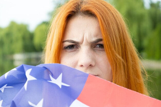 Portrait d'une fille aux cheveux rouges en colère cachant son visage derrière le drapeau national des États-Unis.